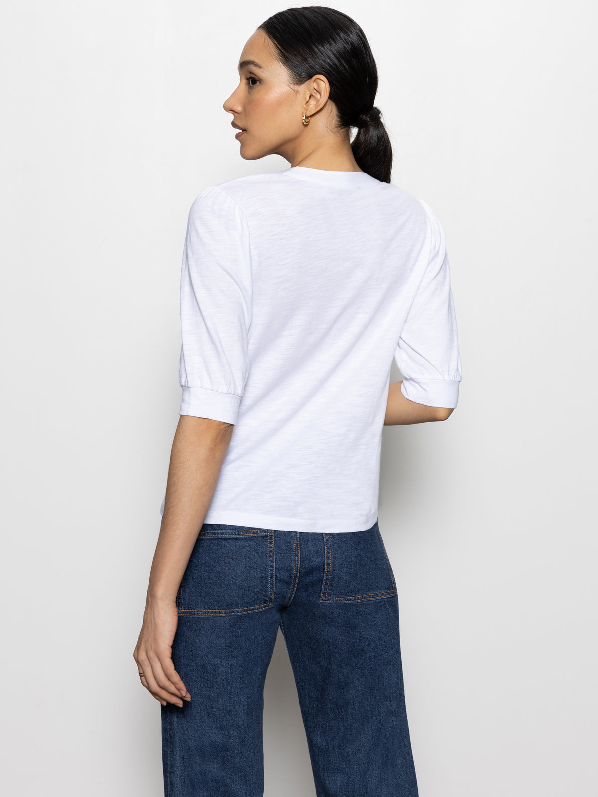 A woman with long dark hair in a low ponytail, wearing the Sanctuary Clothing mixed media tee in white and blue jeans, stands with her back slightly turned to the camera on a plain white background.