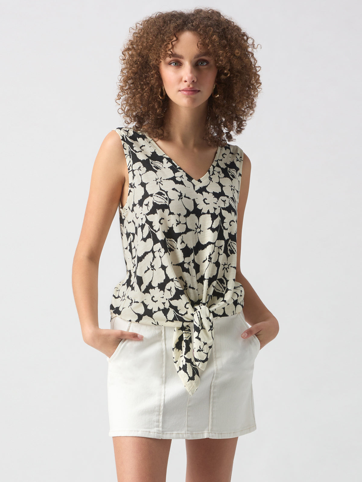 A person with curly hair is wearing the V Neck Tie Tank Echo Blooms by Sanctuary Clothing, featuring a black and white floral pattern and tied at the front. They are also wearing a light-colored skirt with their hands in the pockets. They are standing against a plain background.