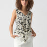 A person with curly hair is wearing the V Neck Tie Tank Echo Blooms by Sanctuary Clothing, featuring a black and white floral pattern and tied at the front. They are also wearing a light-colored skirt with their hands in the pockets. They are standing against a plain background.