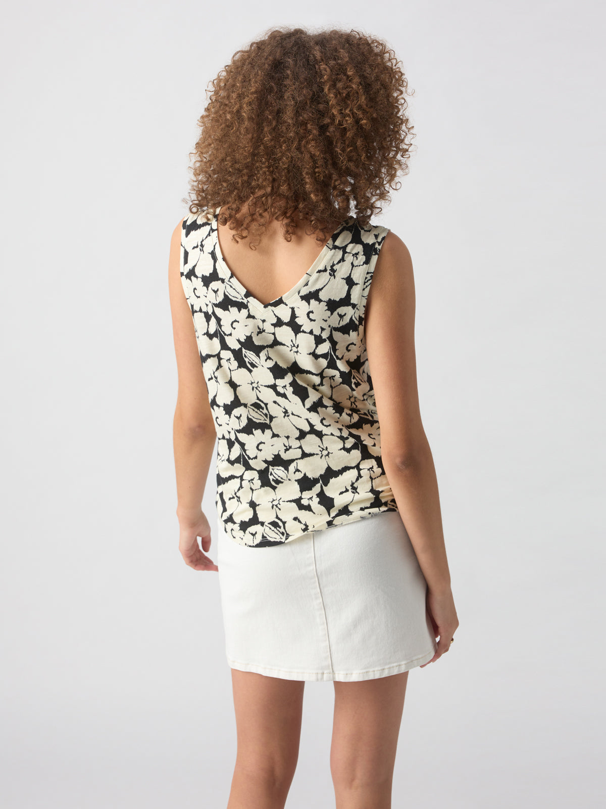 A person with curly hair stands facing away, wearing the V Neck Tie Tank Echo Blooms by Sanctuary Clothing, paired with a white skirt. The background is plain and light grey.