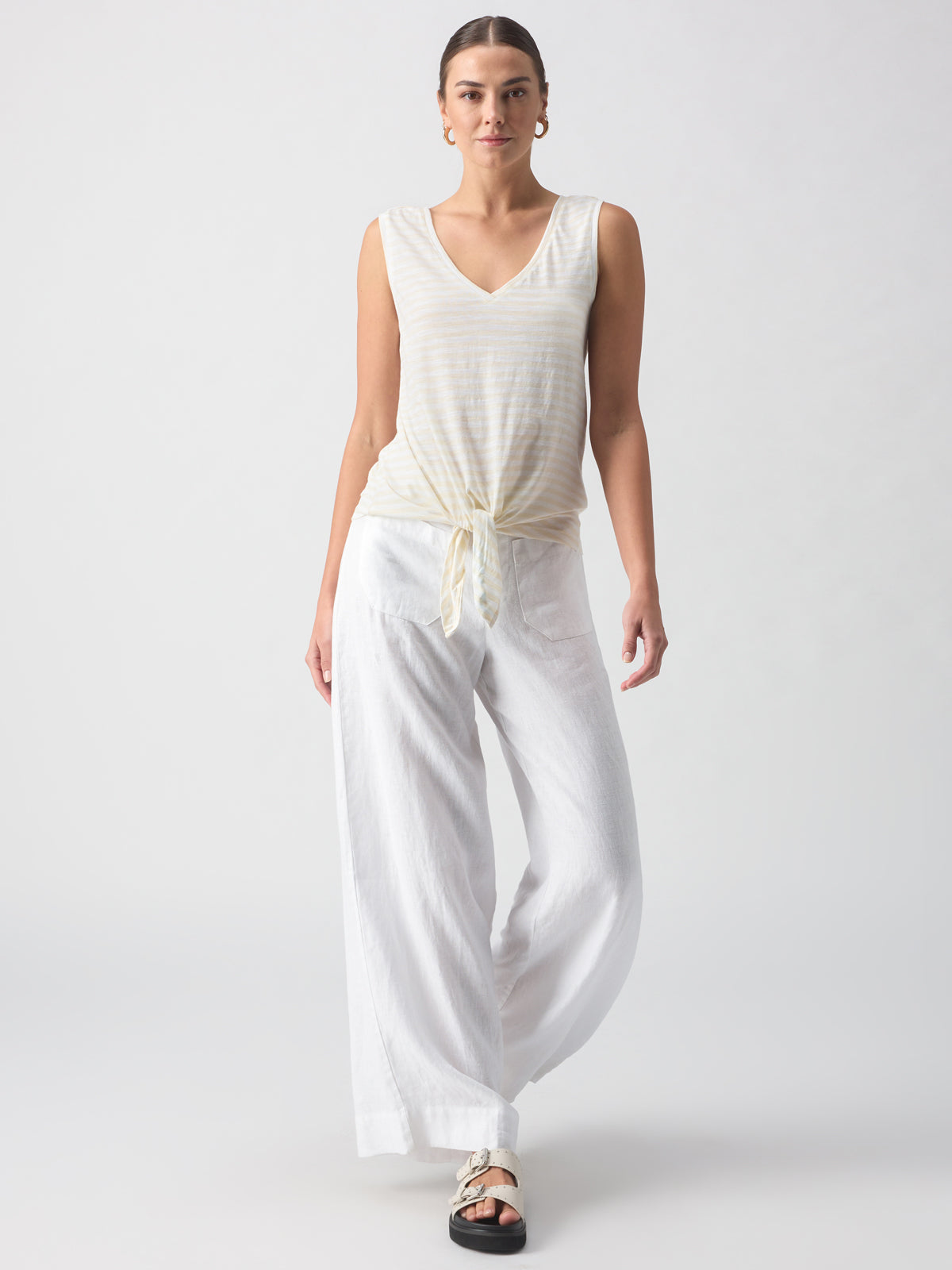 A person wears a white V Neck Tie Tank White Birch Stripe from Sanctuary Clothing with white wide-legged pants. They are also wearing black sandals and large hoop earrings, walking forward against a neutral background.