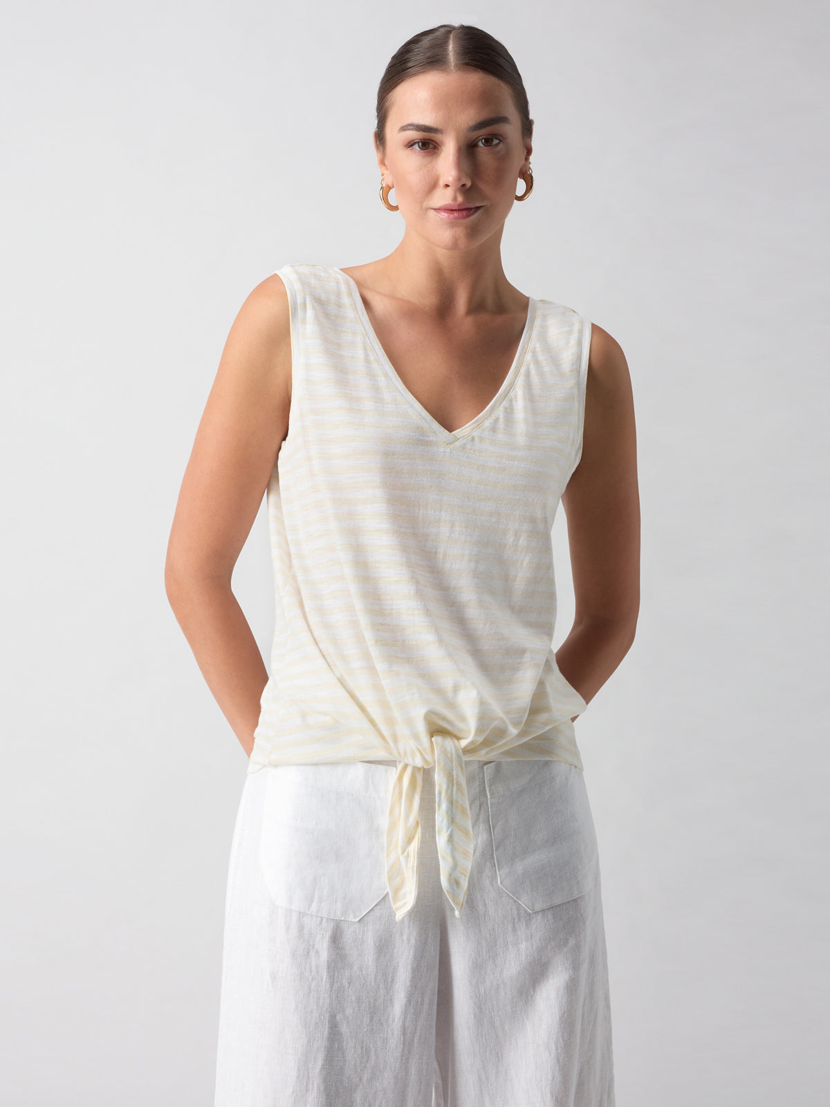 A person with hair tied back wears the V Neck Tie Tank White Birch Stripe from Sanctuary Clothing, a sleeveless, light-colored top with subtle vertical stripes and knotted at the waist. They also wear white pants with their hands tucked into the pockets. The background is a plain, light gray color.