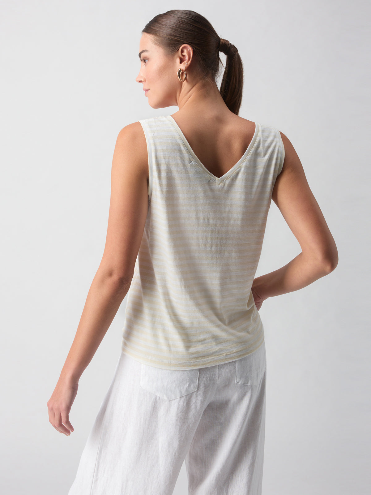 A woman with brown hair tied back in a ponytail is standing facing away. She is wearing the V Neck Tie Tank White Birch Stripe by Sanctuary Clothing and white pants. One hand is resting on her hip, and the background is plain and light gray.