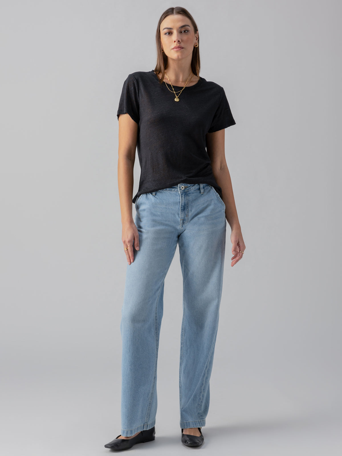 A woman stands against a plain background, wearing the Linen Perfect Tee Black from Sanctuary Clothing, light blue jeans, and black shoes. She has a serious expression and her left arm is relaxed by her side while her right arm rests on her hip. She is accessorized with a necklace and earrings.