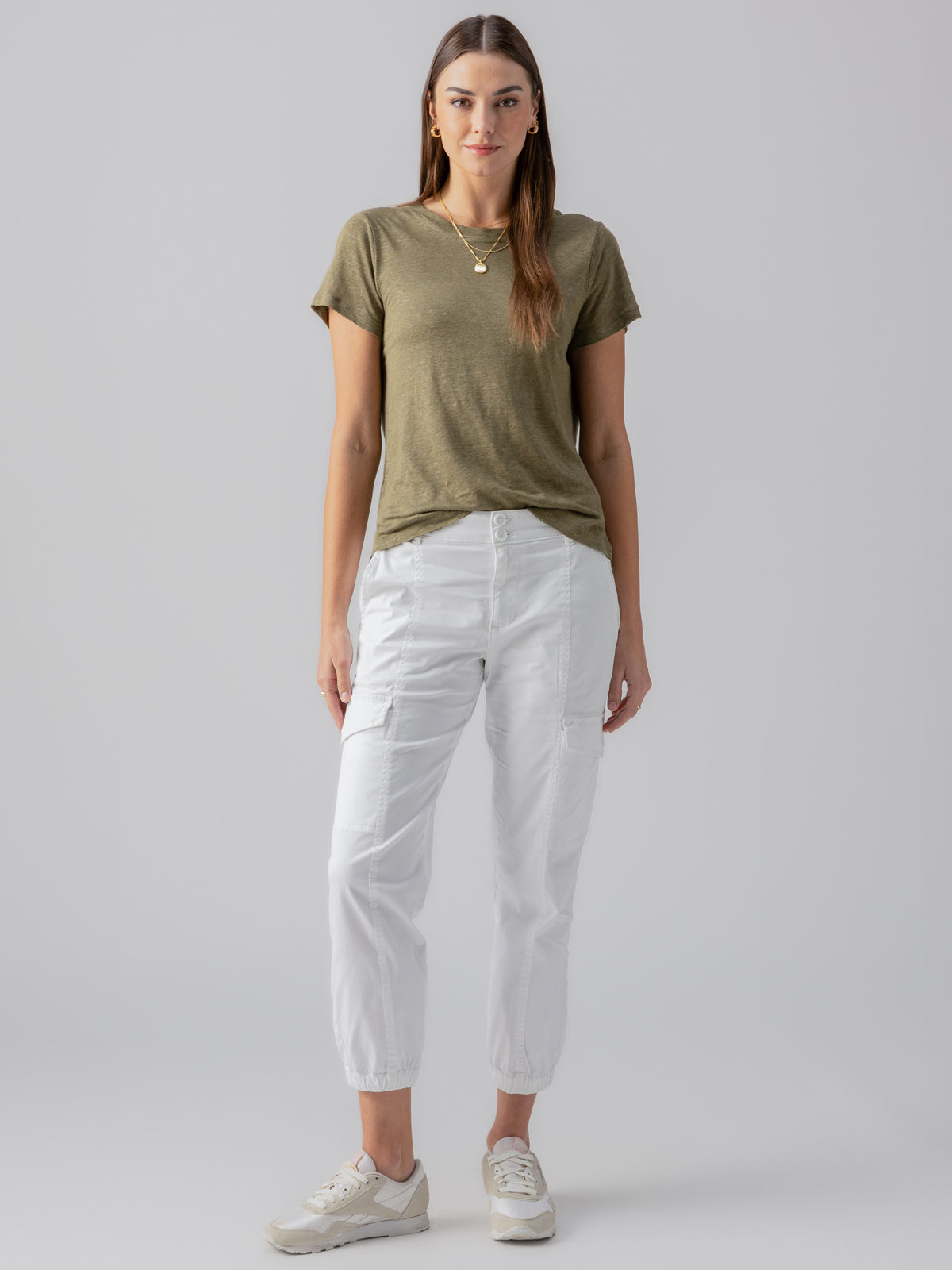 A person with long hair is standing against a plain grey background. They are wearing the Linen Perfect Tee in Burnt Olive by Sanctuary Clothing, white jogger pants, and white sneakers. They have a neutral expression and are accessorized with a gold necklace and earrings.