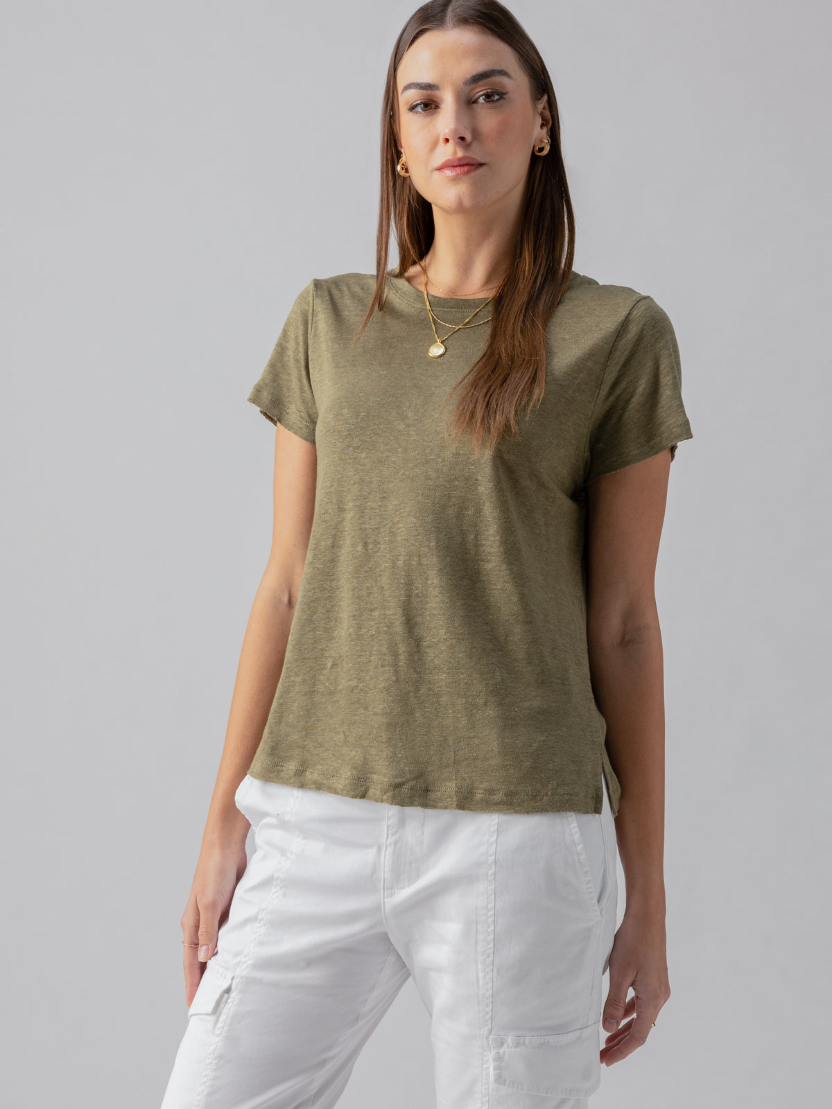 A woman with long brown hair stands against a plain background, wearing the Sanctuary Clothing Linen Perfect Tee in Burnt Olive and white pants. She accessorizes with gold hoop earrings and a gold necklace with a pendant. Her expression is neutral.