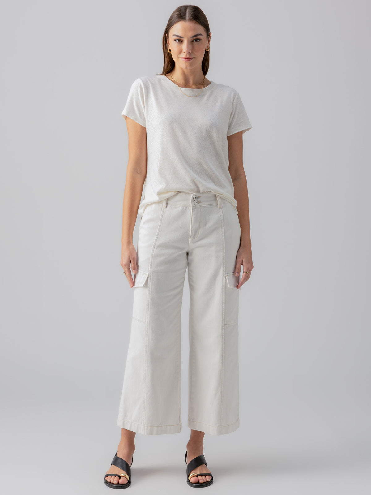 A woman wearing the Sanctuary Clothing Linen Perfect Tee in white and white wide-leg pants with pockets stands against a light gray background. She has long hair and is wearing black slide sandals. She poses with her hands by her sides.