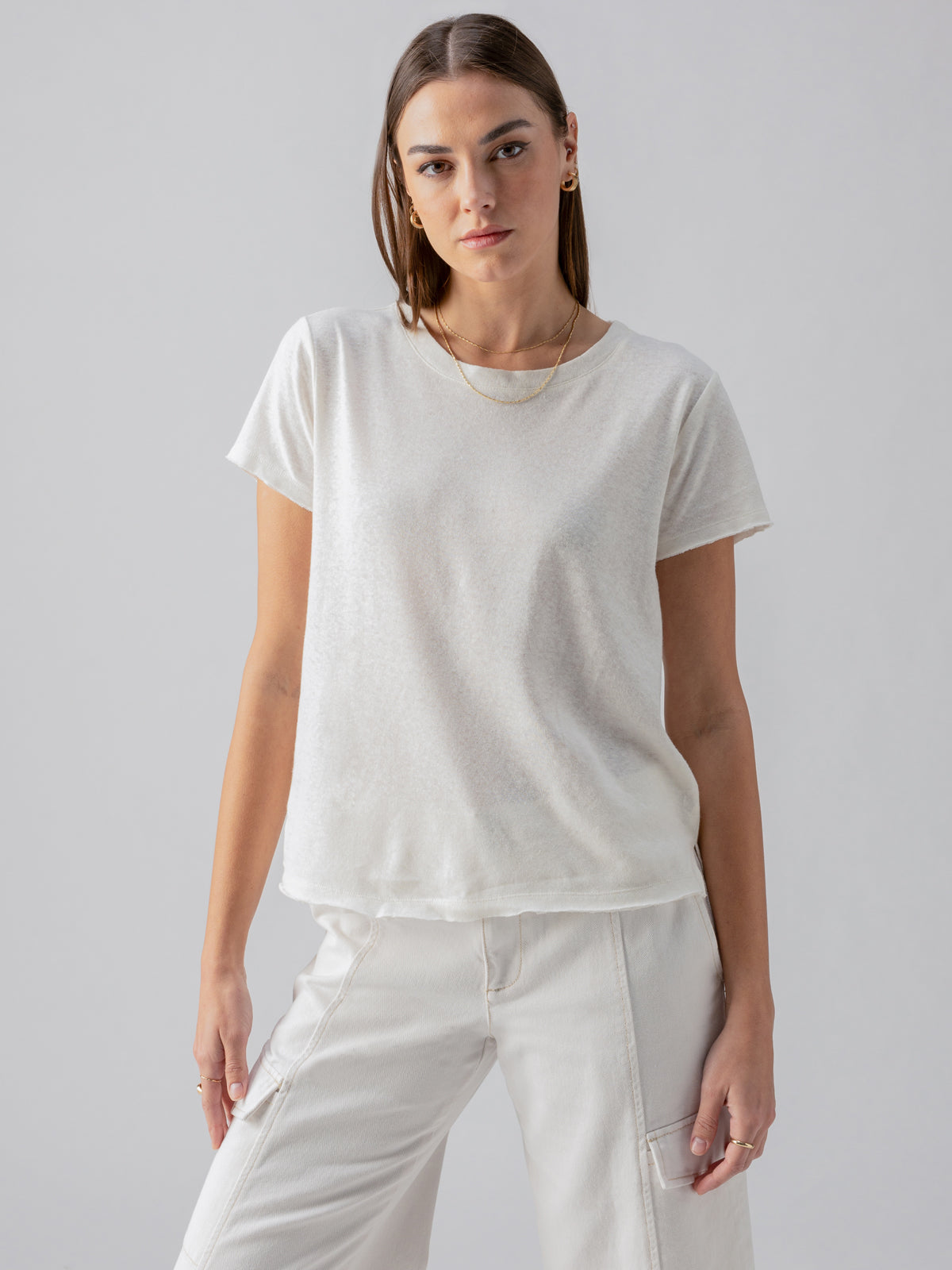 A person with shoulder-length hair is standing against a plain backdrop, wearing the Linen Perfect Tee White by Sanctuary Clothing paired with white pants. They have a neutral expression, and their hands are by their sides. They are also wearing small hoop earrings and a delicate chain necklace.