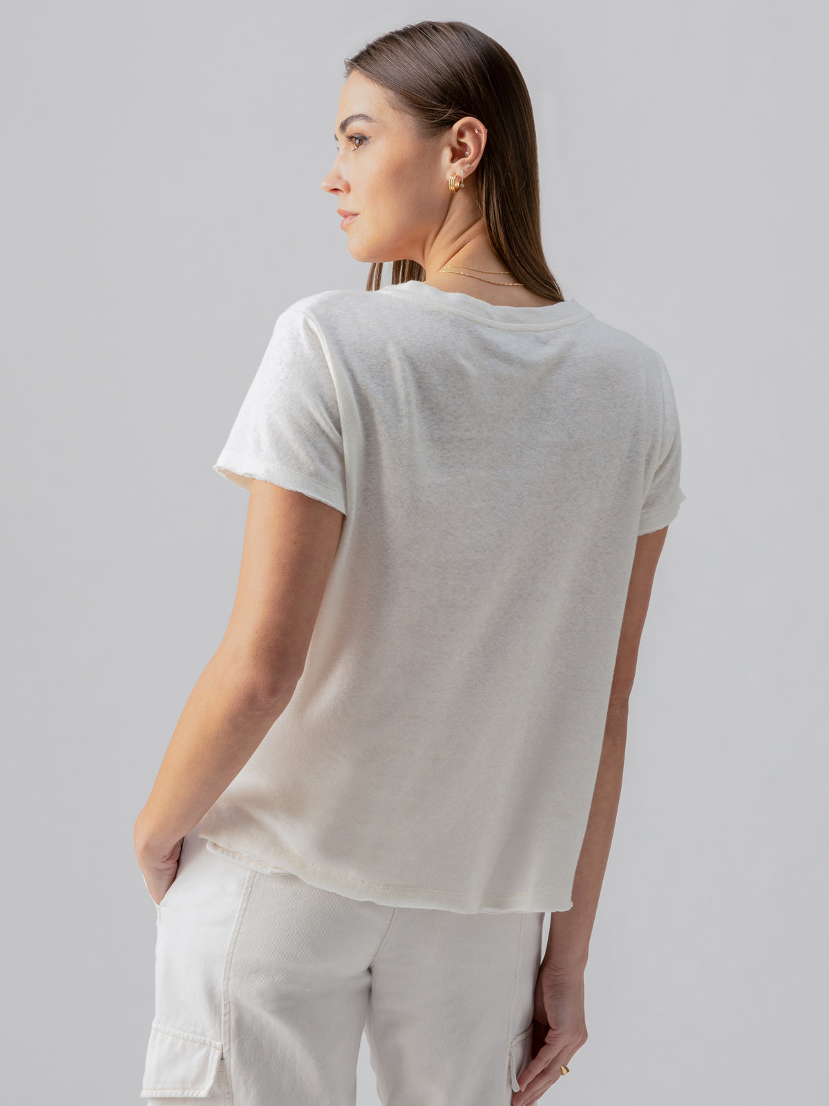 A woman with long, straight brown hair is standing and looking to the side. She is wearing the Linen Perfect Tee White by Sanctuary Clothing along with white pants. Her left hand is tucked into her pocket and the background is plain gray.