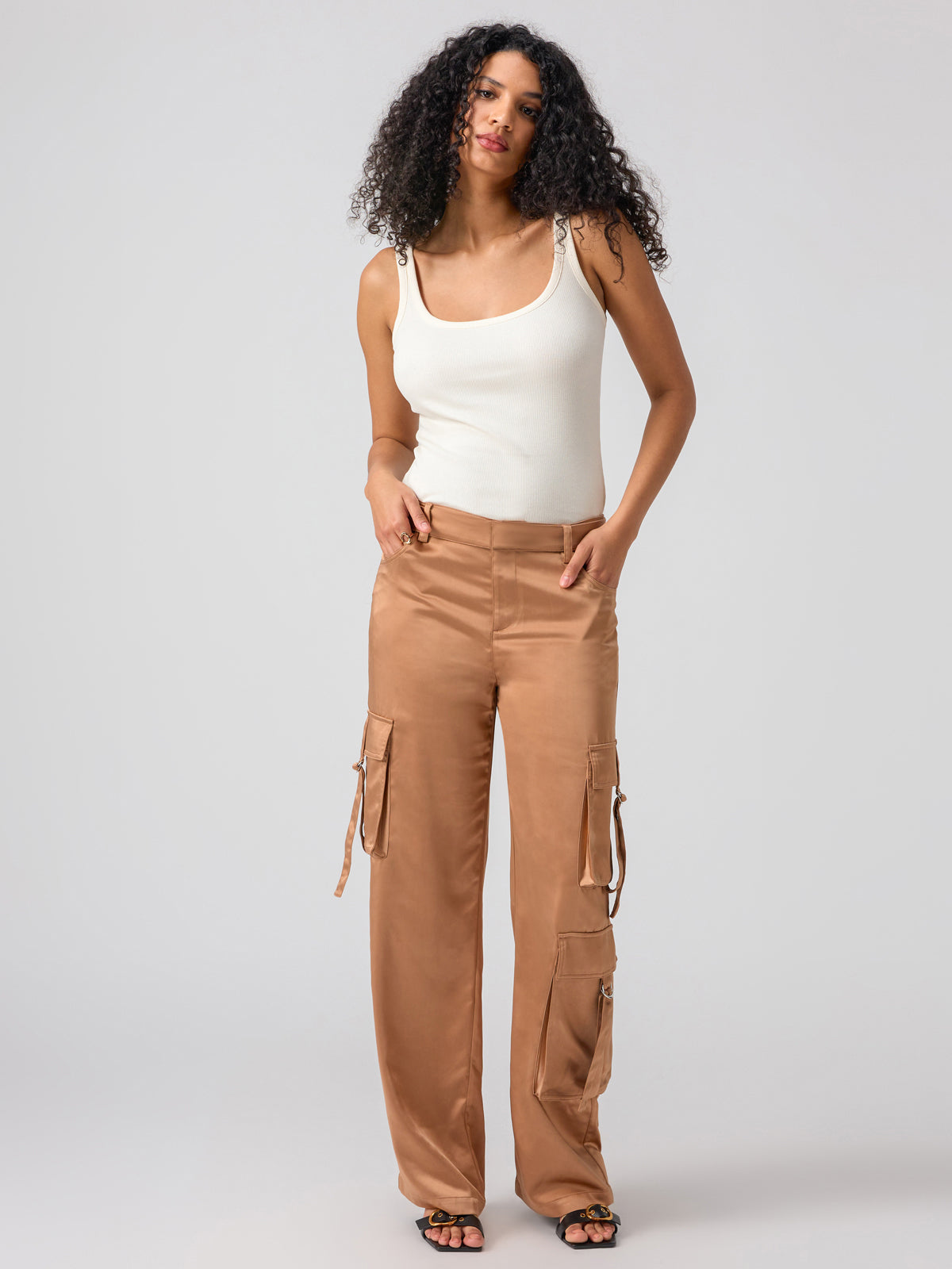 A person with curly hair wearing the My Kind Of Tank Birch by Sanctuary Clothing and tan cargo pants stands against a plain gray background. They have their hands in the pockets of their pants and are looking at the camera with a slight smile.