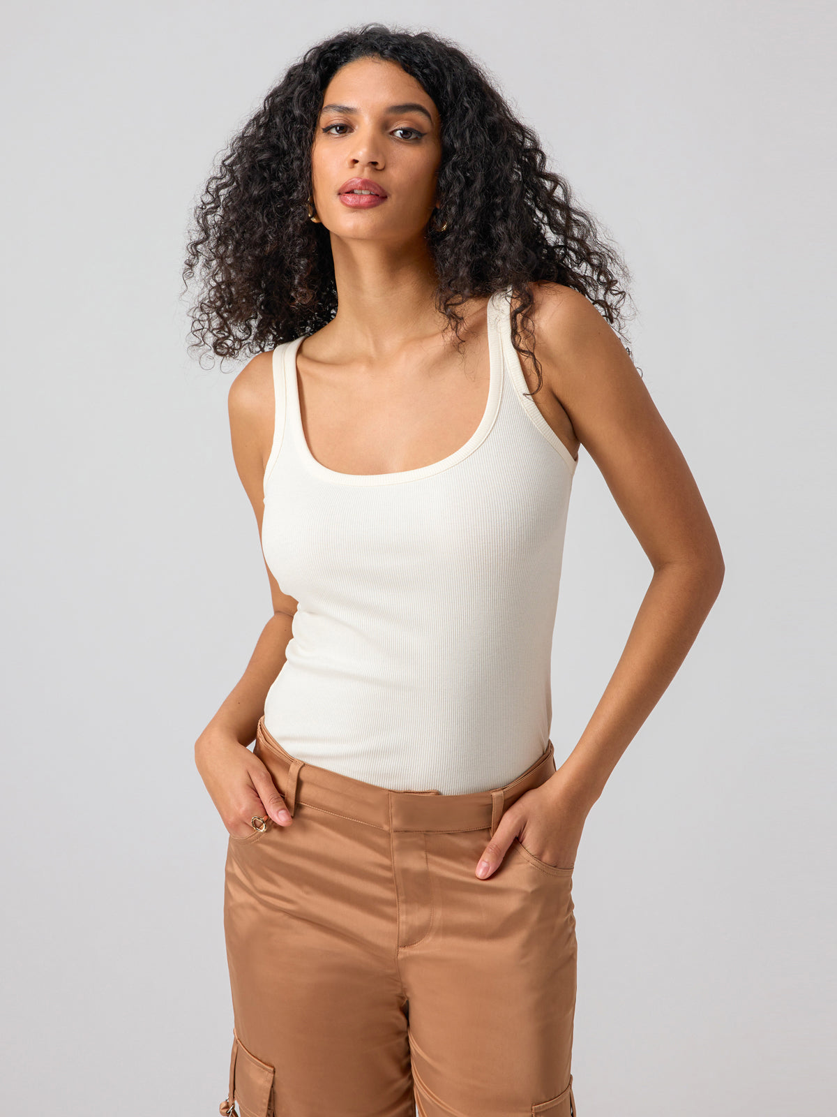 A woman with long, curly hair is posing against a plain background. She is wearing the My Kind Of Tank Birch by Sanctuary Clothing and brown pants. Her hands are in her pockets, and she is looking confidently at the camera.