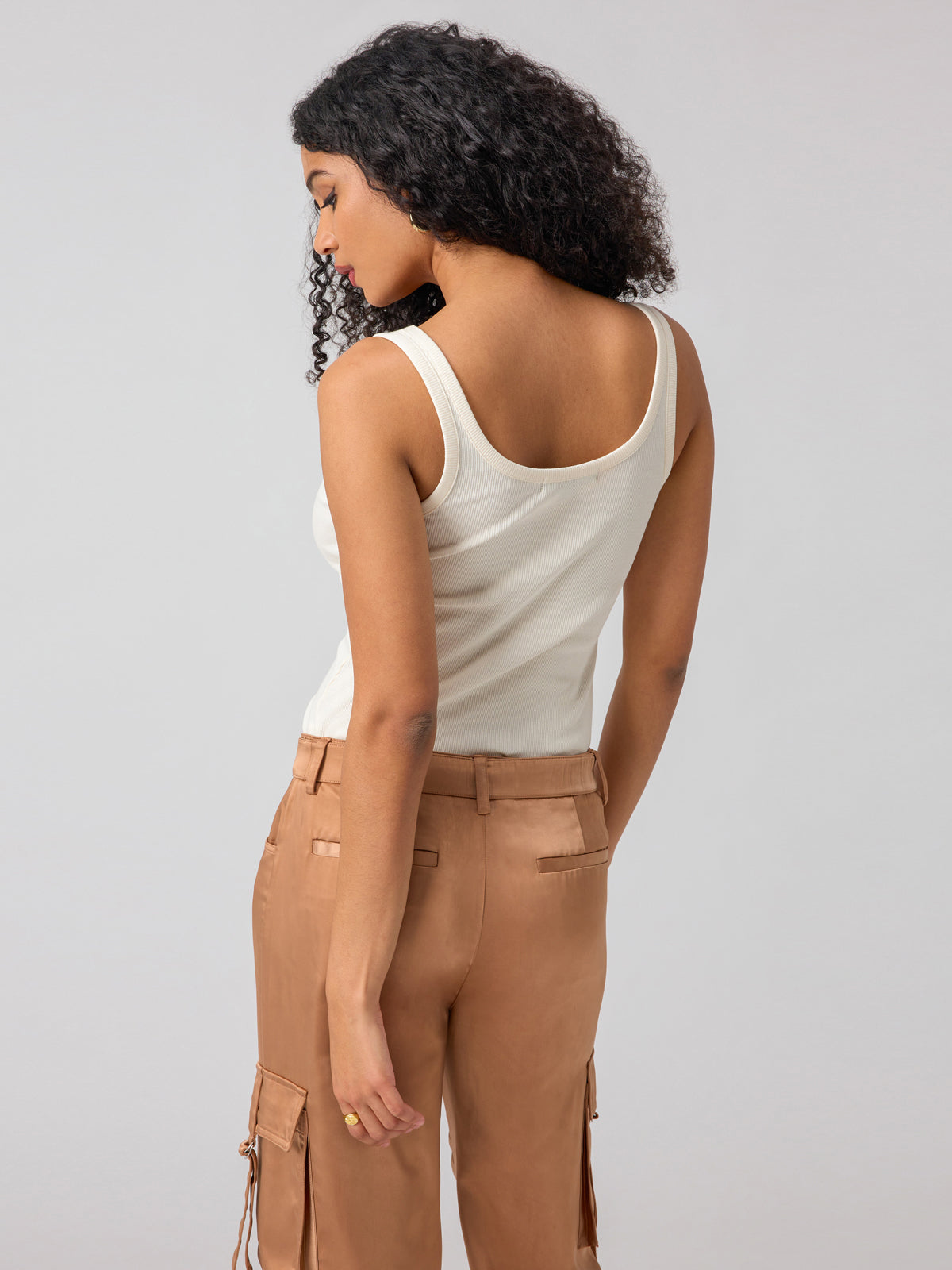 A person with curly hair is seen from the back, wearing the My Kind Of Tank Birch from Sanctuary Clothing. They are also dressed in light brown pants featuring flap pockets and side ties, standing against a plain grey background.