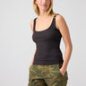 A woman with long blonde hair is wearing the My Kind Of Tank Black from Sanctuary Clothing and camouflage-patterned shorts. She has her hands in her pockets and is standing against a plain gray background.