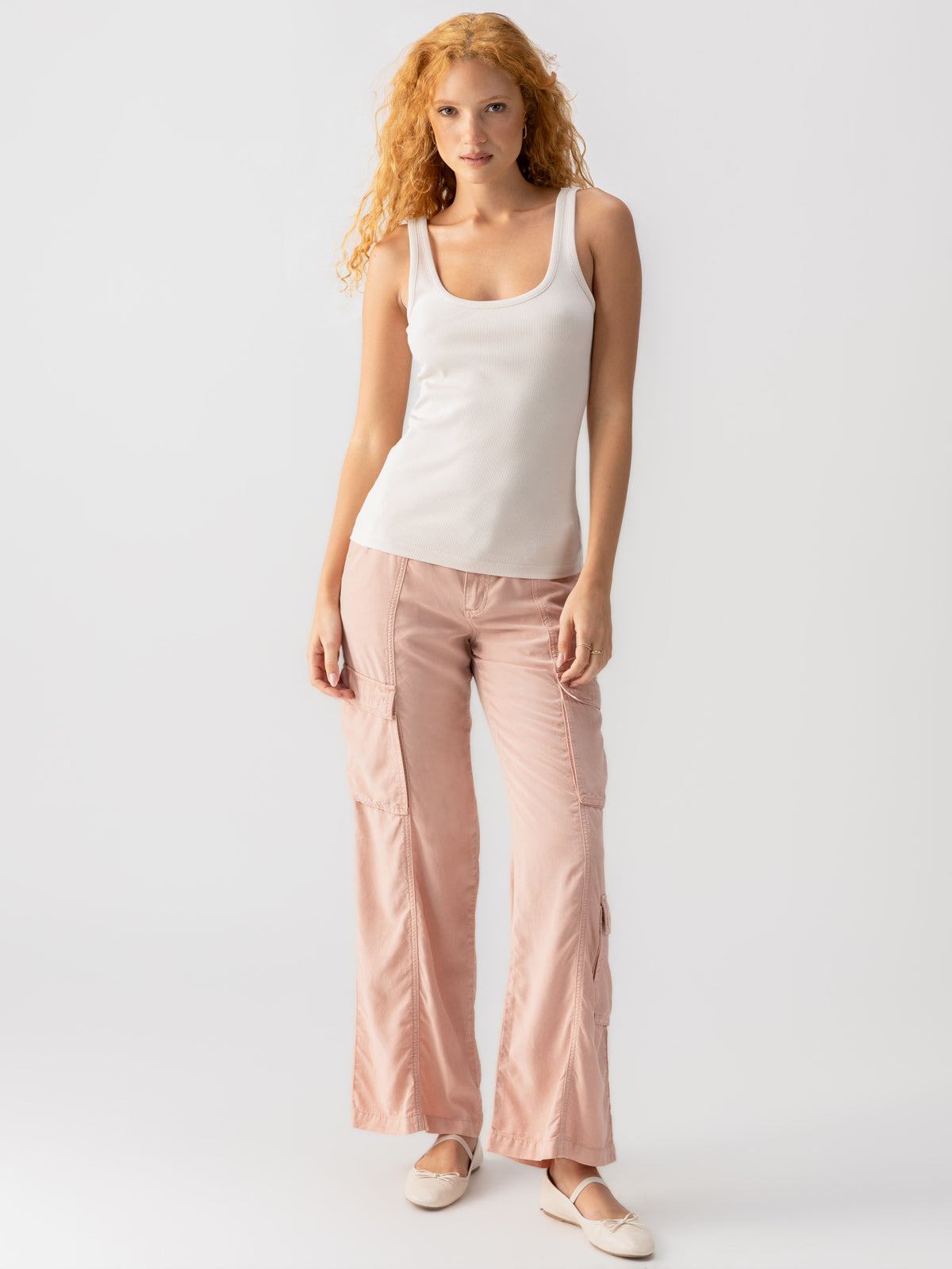 A person with long, curly hair is standing against a plain background. They are wearing the Sanctuary Clothing My Kind Of Tank Crystal Stone, light pink cargo-style pants with multiple pockets, and beige ballet flats. The person is looking straight ahead with a relaxed expression.