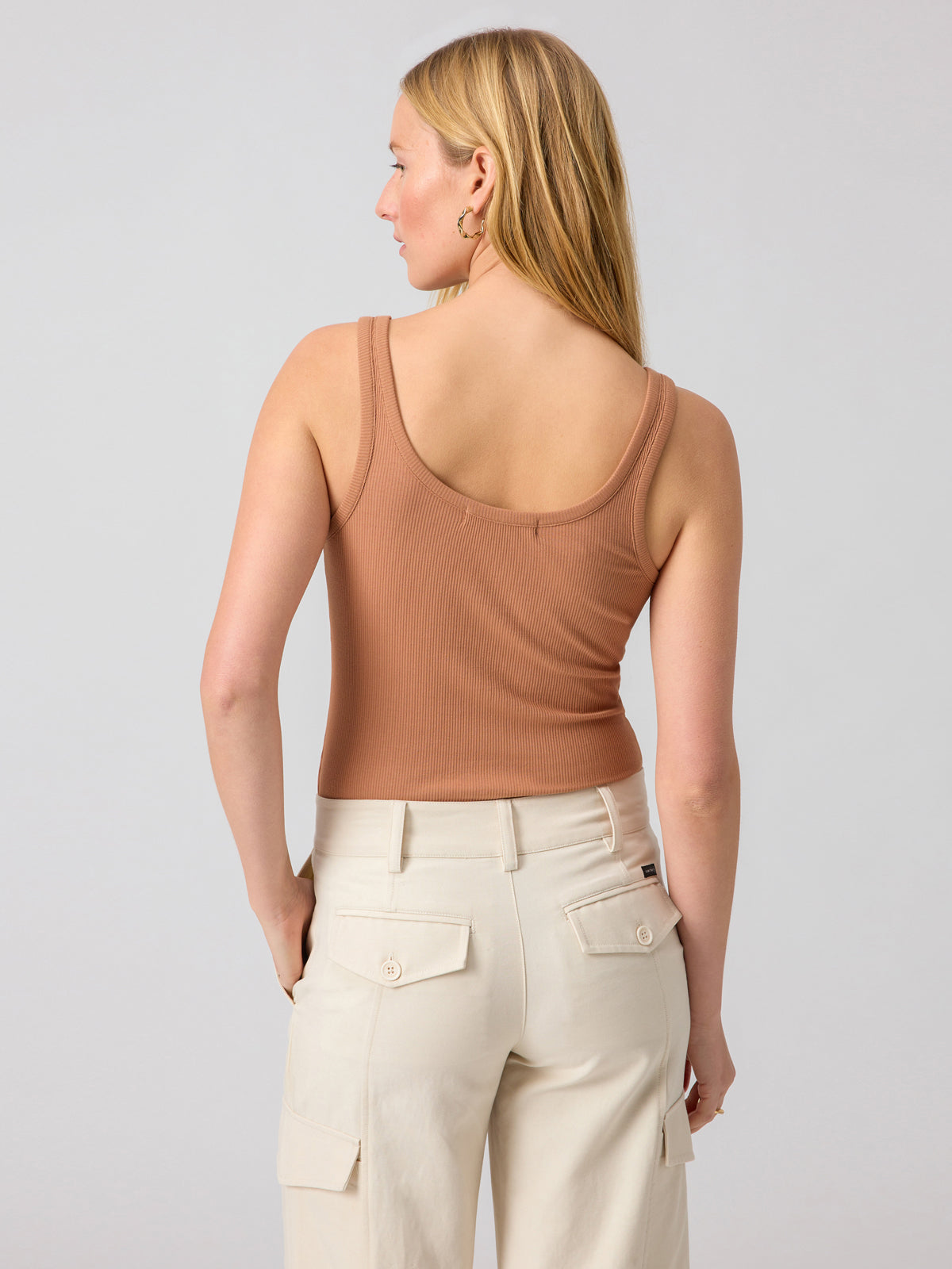 A person with long blonde hair stands facing away from the camera, donning the Sanctuary Clothing's 'My Kind Of Tank' in Mocha Mousse and light beige cargo pants with multiple pockets. The background is plain and light-colored.