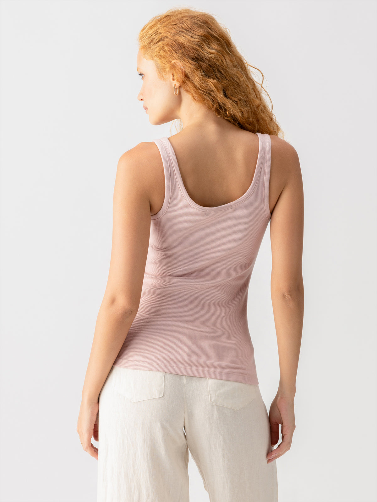 A person with long, curly hair is shown from the back against a plain white background, wearing the My Kind Of Tank in Smokey Rose by Sanctuary Clothing and white pants. The person is facing slightly to the left with a relaxed posture.