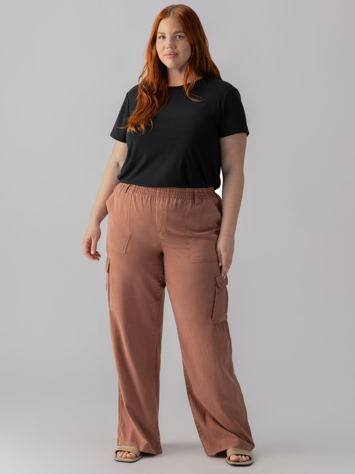 A person with long red hair stands against a plain gray background. They are wearing the Perfect Mesh Tee Black from Sanctuary Clothing's Inclusive Collection and loose-fitting, high-waisted, brown cargo pants. Their hands are relaxed by their sides, and they are looking directly at the camera.