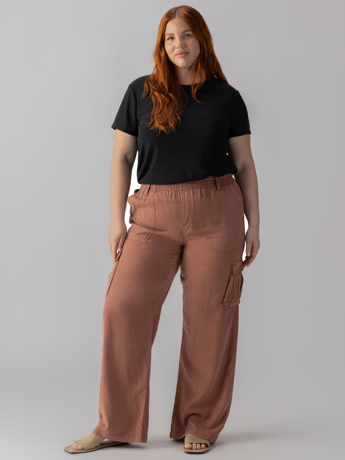 A woman with long red hair is wearing the Relaxed Reissue Washed Clay Inclusive Collection from Sanctuary Clothing, which includes a black t-shirt, light brown cargo pants, and beige sandals. She stands with a relaxed posture against a plain grey background, with one hand on her hip and the other hanging by her side.