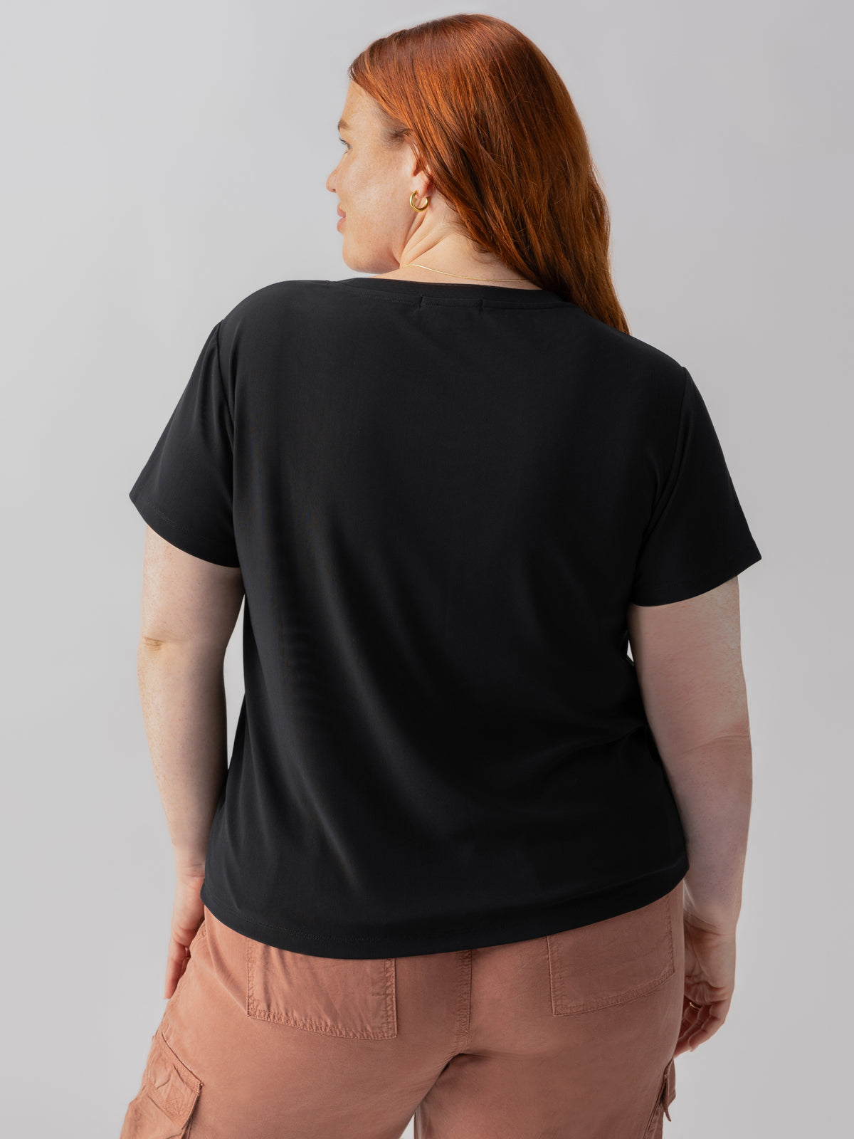 A person with long red hair, seen from the back, is wearing the "Perfect Mesh Tee Black" from Sanctuary Clothing's Inclusive Collection, along with light brown pants that feature side pockets. The background is a plain grey color.