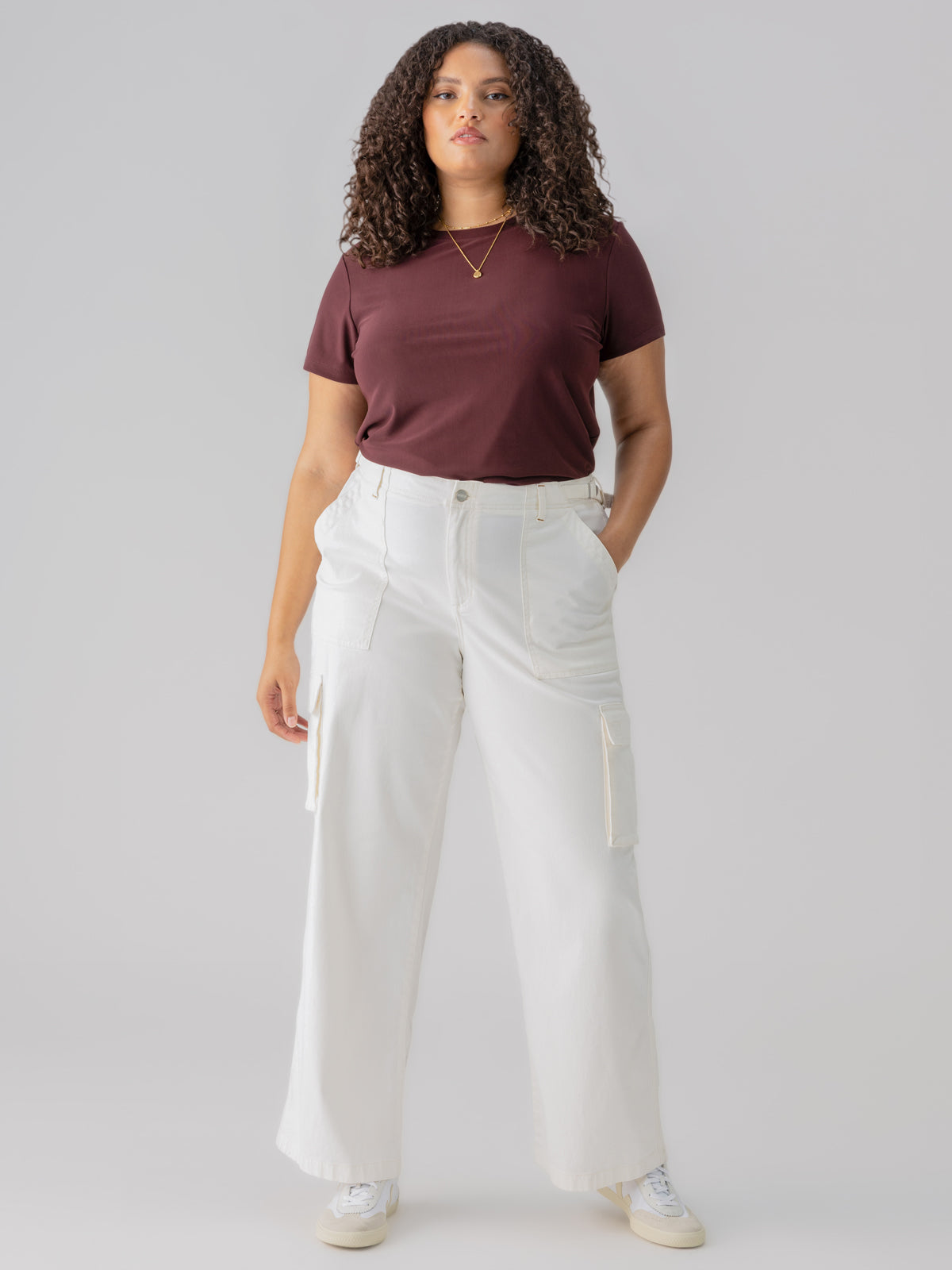 A person with curly hair is wearing the "Perfect Mesh Tee Dark Cherry Inclusive Collection" by Sanctuary Clothing, along with white high-waisted cargo pants, their hands placed in the pockets. They are standing against a plain grey background and are wearing white sneakers.