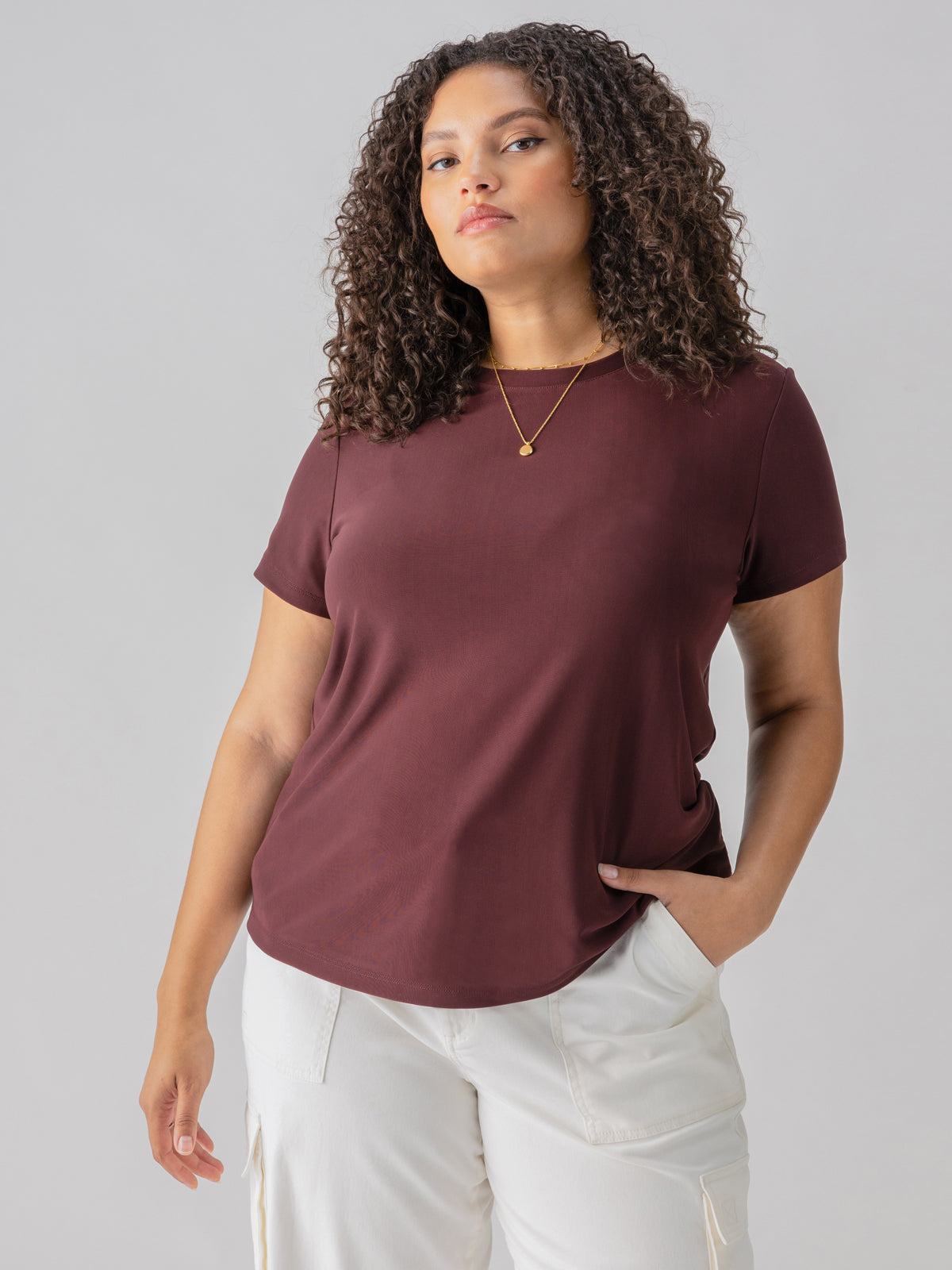 A woman wears the Sanctuary Clothing Perfect Mesh Tee from the Inclusive Collection in Dark Cherry and white pants, standing against a neutral background. She has curly hair and is accessorized with a simple necklace. Her left hand is in her pocket as she gazes forward with a serious expression.