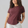 A woman wears the Sanctuary Clothing Perfect Mesh Tee from the Inclusive Collection in Dark Cherry and white pants, standing against a neutral background. She has curly hair and is accessorized with a simple necklace. Her left hand is in her pocket as she gazes forward with a serious expression.