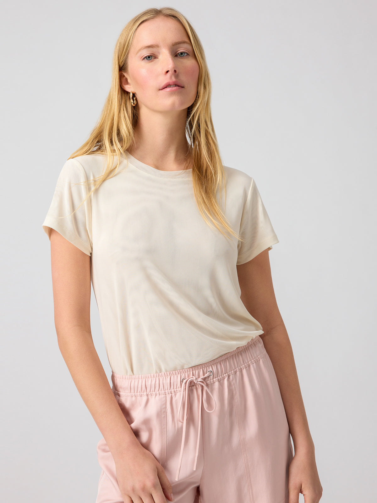 A woman with long blonde hair is wearing Sanctuary Clothing's Perfect Mesh Tee Birch and light pink drawstring pants. She stands against a plain, light-colored background and looks forward with a neutral expression.