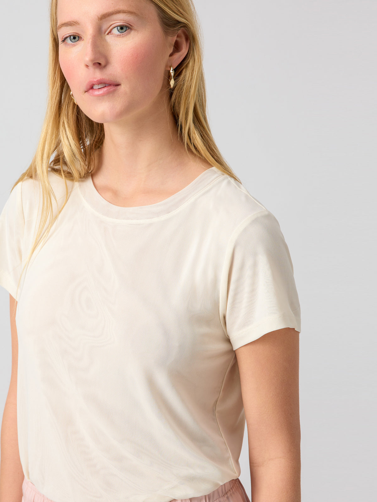 A woman with long blonde hair is wearing a light beige Perfect Mesh Tee Birch short-sleeve blouse from Sanctuary Clothing and gold hoop earrings. She is gazing slightly to the side against a plain, light gray background. Her expression is neutral.