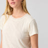 A woman with long blonde hair is wearing a light beige Perfect Mesh Tee Birch short-sleeve blouse from Sanctuary Clothing and gold hoop earrings. She is gazing slightly to the side against a plain, light gray background. Her expression is neutral.