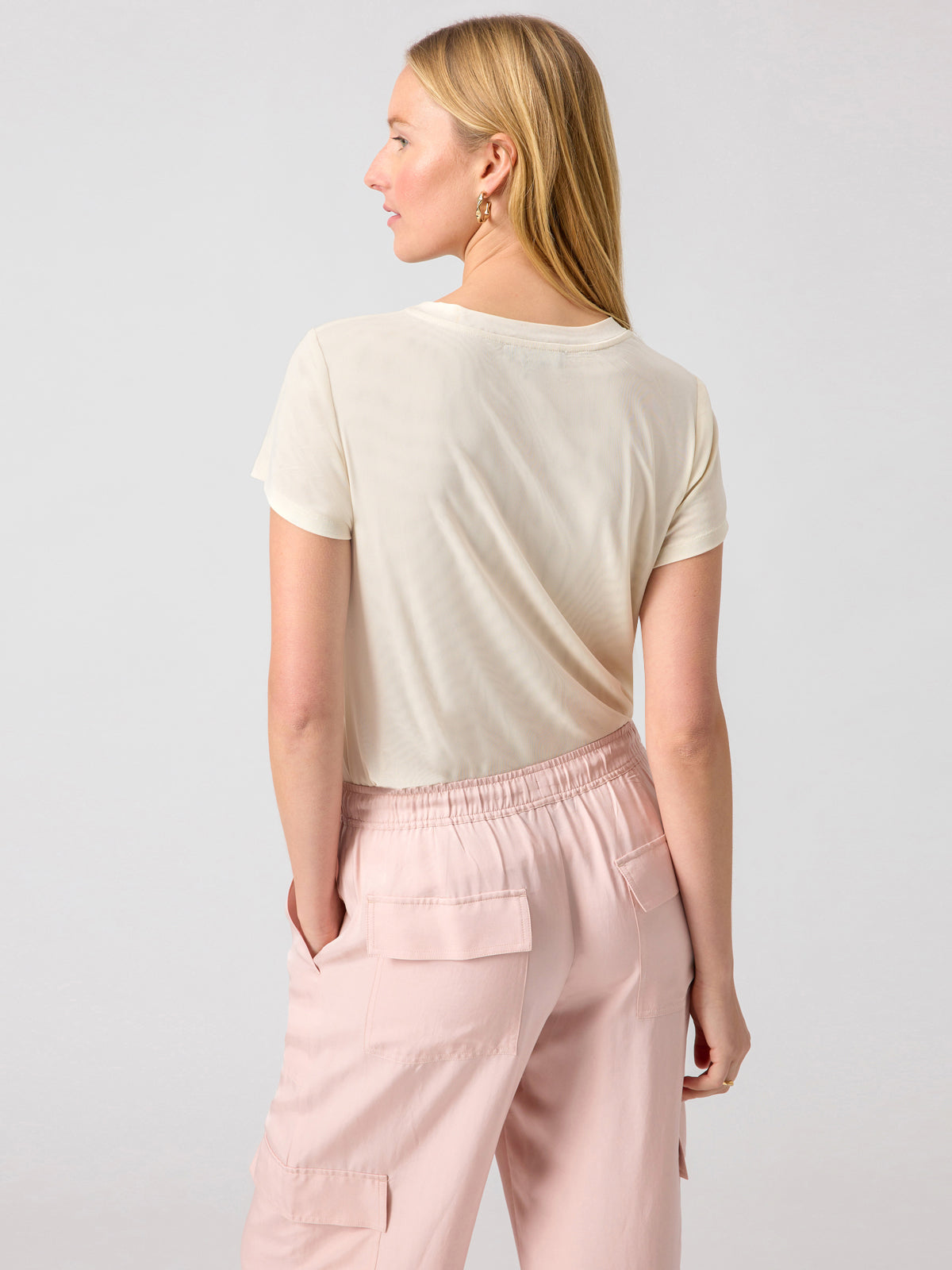 A person with long blonde hair is standing with their back to the camera. They are wearing a light-colored short-sleeve top, specifically the Perfect Mesh Tee Birch by Sanctuary Clothing, and light pink cargo pants with their hands in their pockets. The background is plain and light-colored.