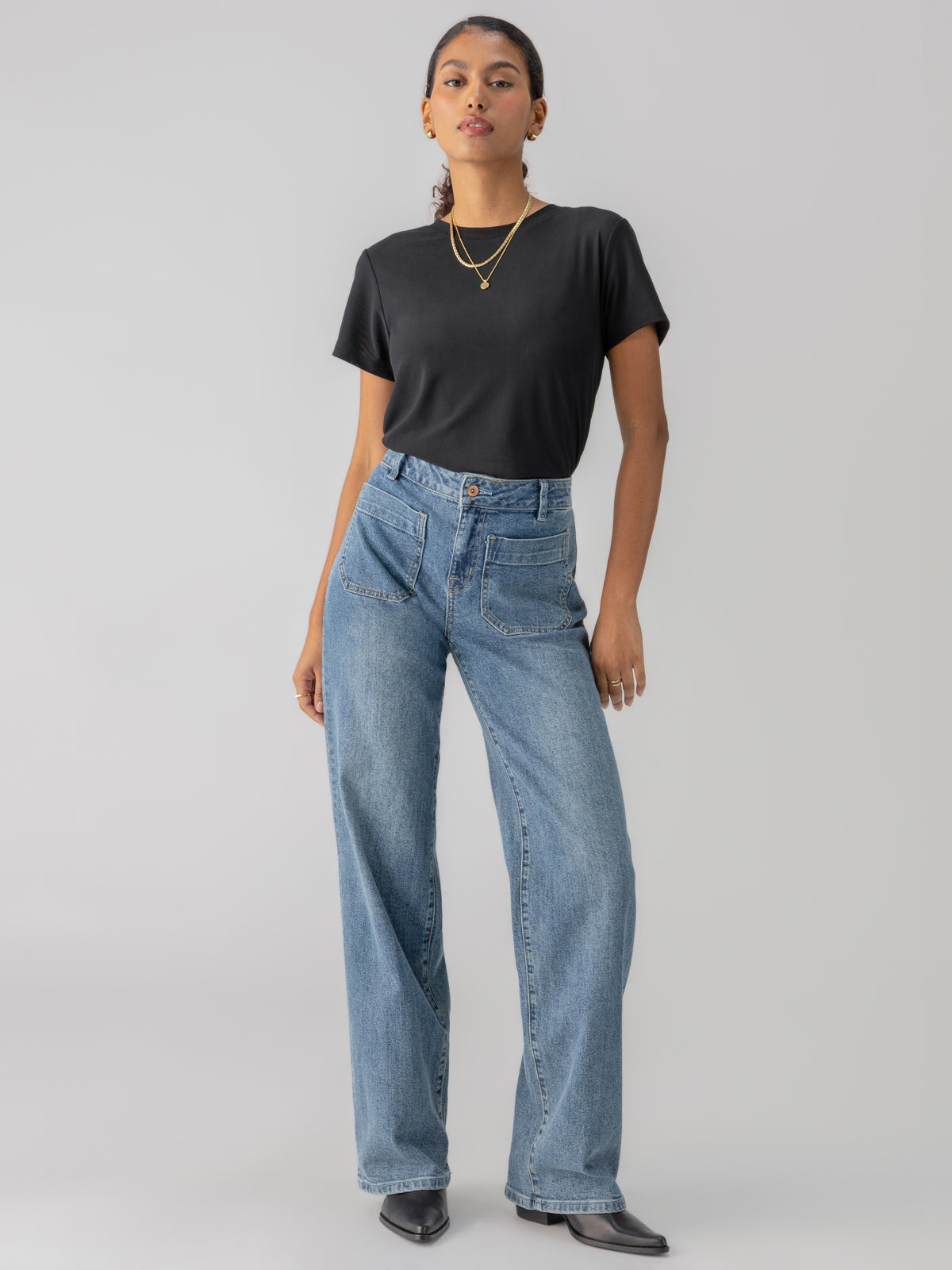 A person with long hair tied back is posing confidently in the Perfect Mesh Tee Black from Sanctuary Clothing, paired with high-waisted blue jeans and black pointed-toe shoes. They are wearing gold hoop earrings and a chain necklace, standing against a plain, light grey background.