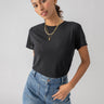 A person with long, dark hair, tied back, wearing the Perfect Mesh Tee Black by Sanctuary Clothing, a pair of blue jeans with front pockets, and gold necklaces. The person stands with one hand on their hip and the other by their side, looking directly at the camera.