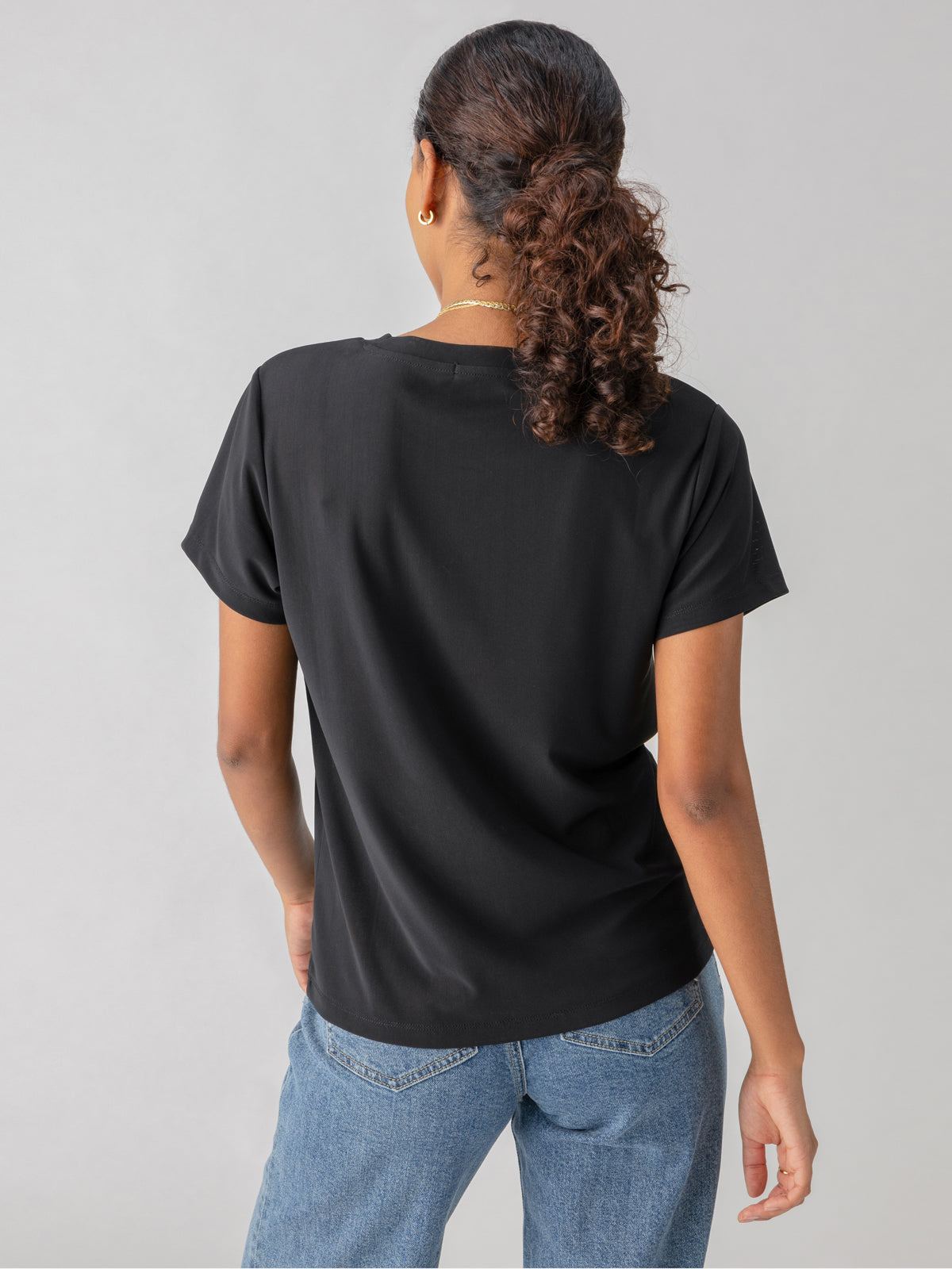 A person with curly hair tied back in a ponytail is seen from behind, wearing the Perfect Mesh Tee Black by Sanctuary Clothing and blue jeans. They stand against a light gray background.