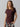 A person with long hair is wearing the Perfect Mesh Tee Dark Cherry by Sanctuary Clothing and patterned pants with abstract, multicolored designs. They are standing against a plain background, smiling softly with one hand in the pocket of their pants.