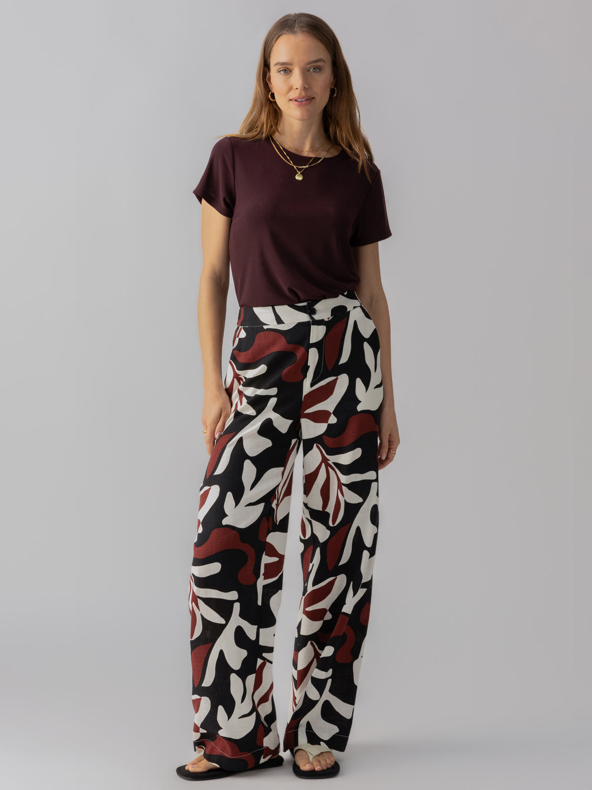 A woman with long hair stands against a plain background, wearing a short-sleeve maroon top and Sanctuary Clothing's "The Soft Trouser Mineral," high-waisted wide-leg pants featuring a bold, abstract leaf pattern in black, white, and red. She accessorizes with layered necklaces and black sandals.