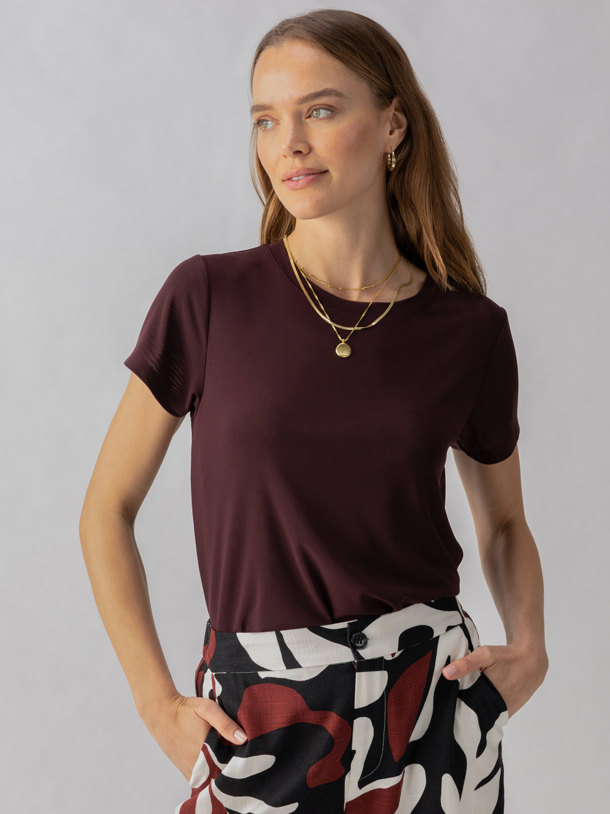 A woman with long, light brown hair stands against a plain background. She is wearing the Perfect Mesh Tee in Dark Cherry by Sanctuary Clothing, layered gold necklaces, and black-and-white patterned pants with red accents. Her hands are in her pockets, and she is looking slightly to the side.