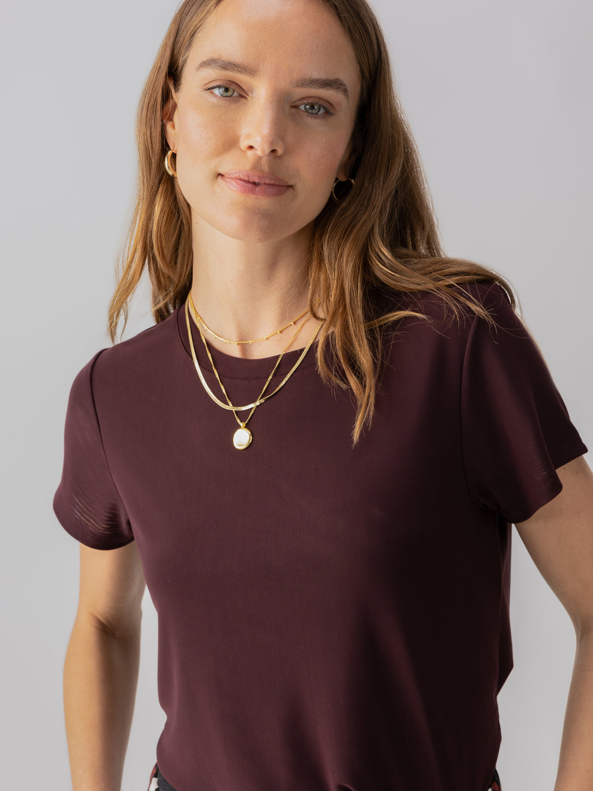 A person with long brown hair wearing the Perfect Mesh Tee Dark Cherry from Sanctuary Clothing and layered gold necklaces, including a pendant, is looking at the camera against a light gray background. They have a slight smile and are accessorized with small gold hoop earrings.