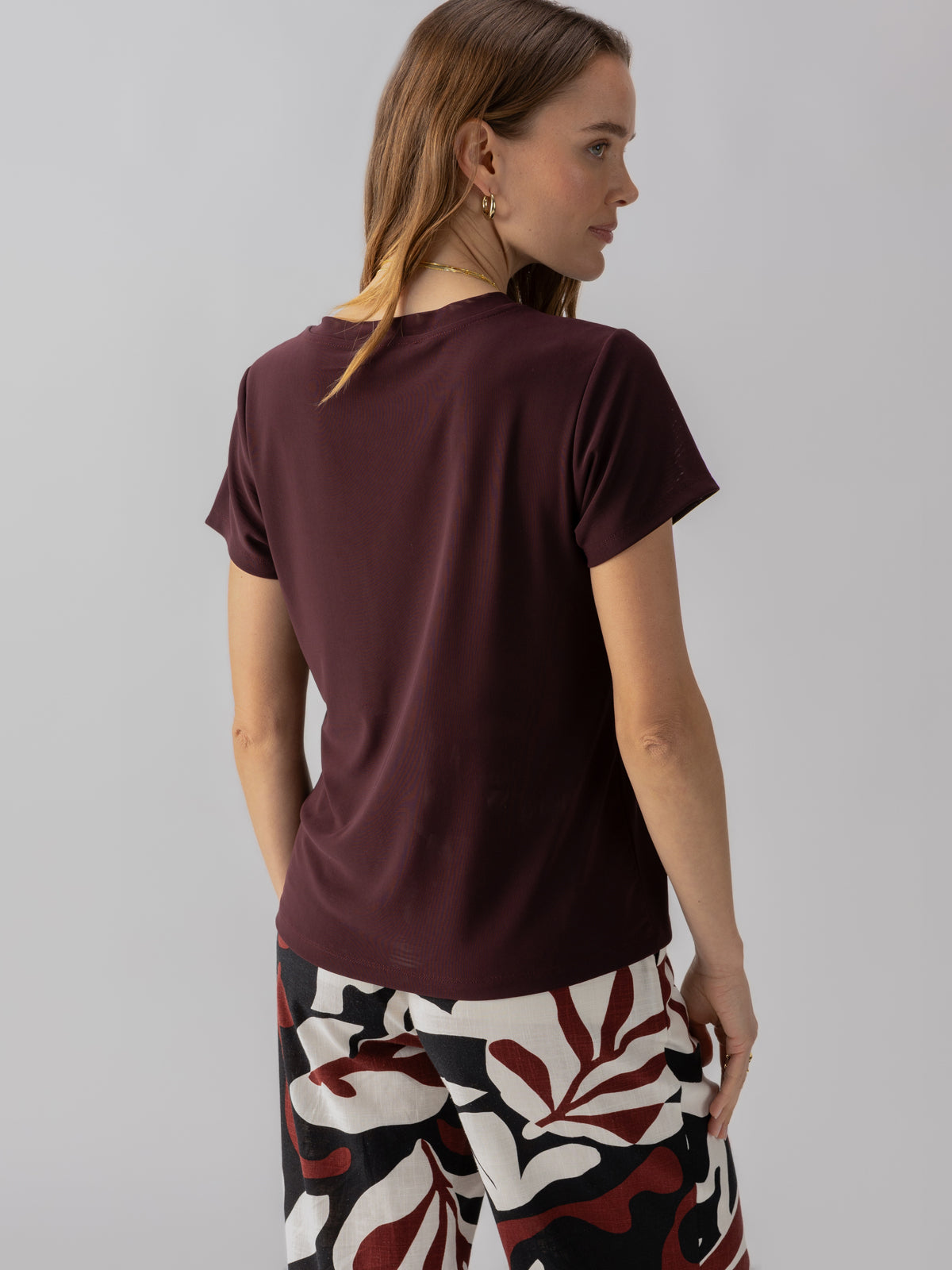 A person with long, light brown hair is shown from the back, wearing the Sanctuary Clothing Perfect Mesh Tee in Dark Cherry and patterned trousers with red, black, and white abstract designs. The person is set against a plain, light gray background.