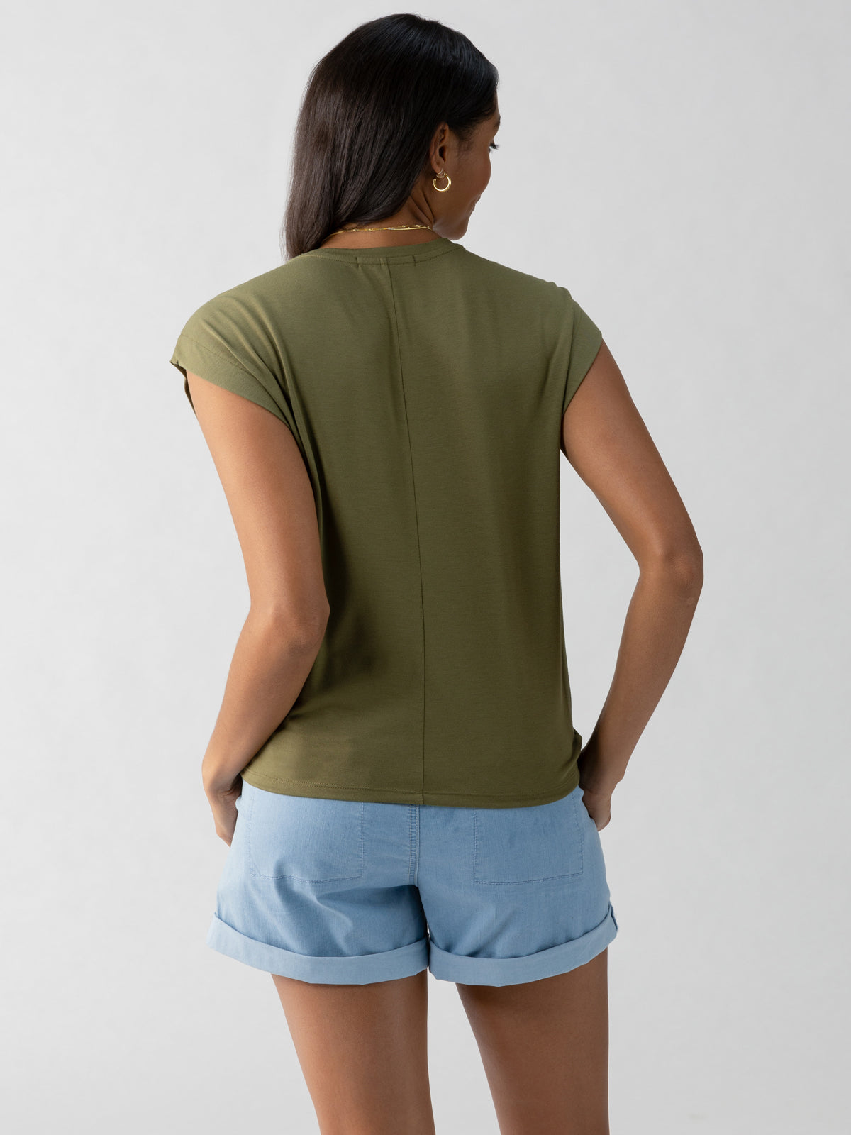 A person with long, dark hair is facing away from the camera. They are wearing a short-sleeve New Era Tee in Burnt Olive from Sanctuary Clothing and light blue denim shorts. The background is plain and light-colored.