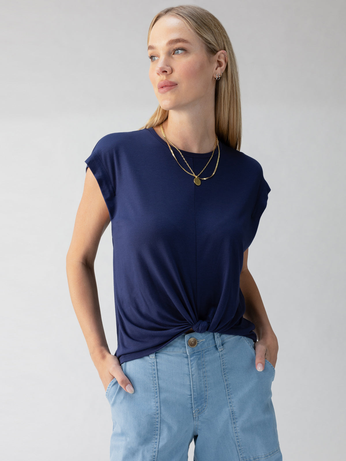 A person with long blonde hair wears Sanctuary Clothing's navy blue New Era Tee Deep Sea, a short-sleeve top with a knotted front, paired with light blue high-waisted jeans. They accessorize with a layered gold necklace featuring a pendant. The person is standing with hands in pockets, looking off to the side.