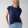 A person with long blonde hair wears Sanctuary Clothing's navy blue New Era Tee Deep Sea, a short-sleeve top with a knotted front, paired with light blue high-waisted jeans. They accessorize with a layered gold necklace featuring a pendant. The person is standing with hands in pockets, looking off to the side.