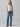 A woman stands against a plain background wearing the New Era Tee White by Sanctuary Clothing, tied at the waist, paired with blue high-waisted straight-leg jeans. She is also sporting black shoes and minimal jewelry. Her long hair cascades down, framing her neutral expression.