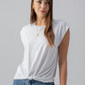 A woman with long, brown hair is wearing the New Era Tee White by Sanctuary Clothing and blue jeans. She accessorizes with layered gold necklaces and hoop earrings. She stands against a plain gray background, looking slightly off-camera.