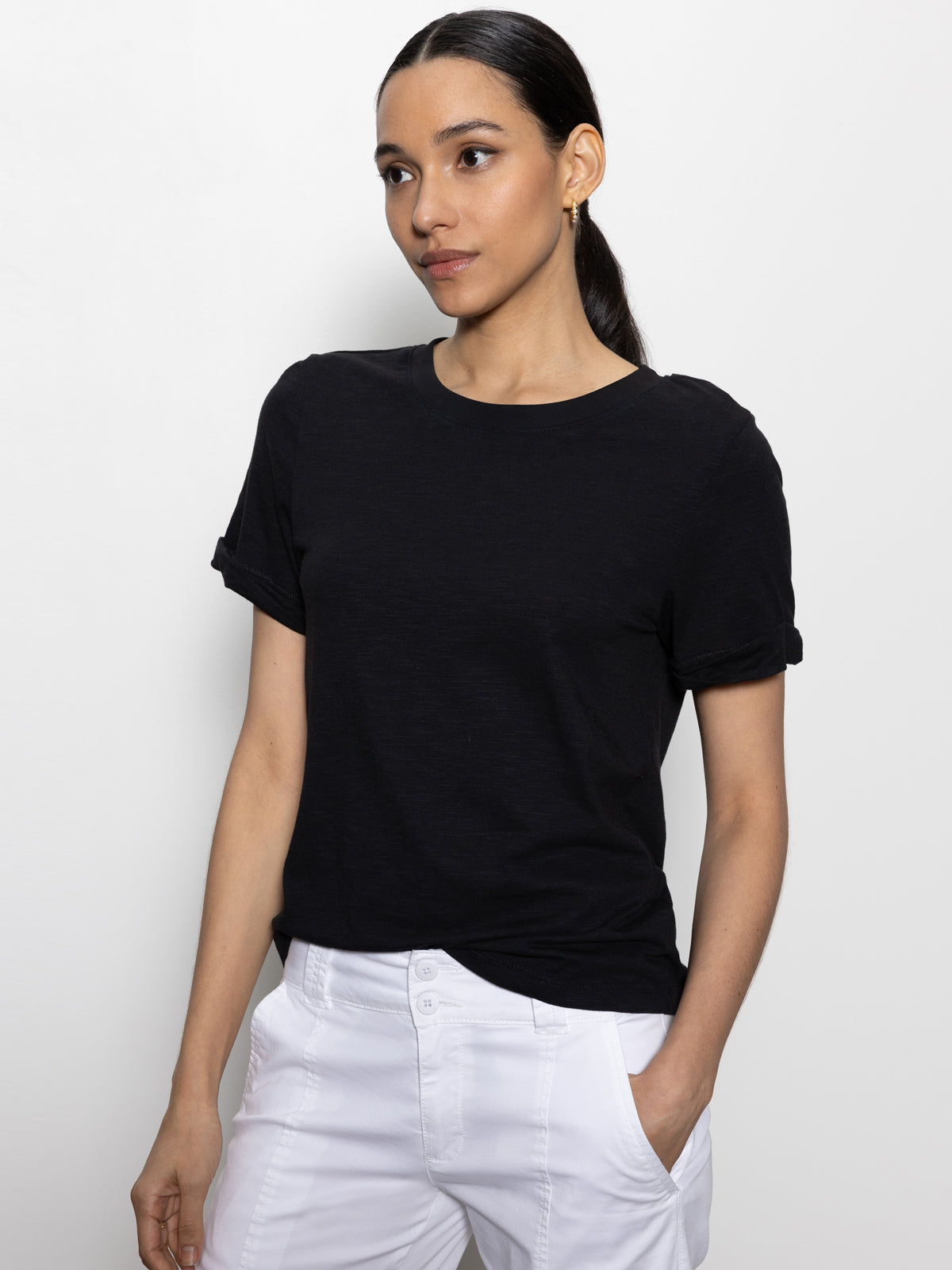 A person with long dark hair in a ponytail wears the Sanctuary Clothing "perfect tee remix black" and white pants, posing against a plain white background with one hand in their pocket, looking to the side.