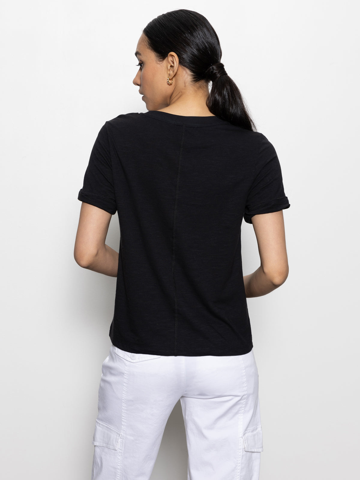 A person with long dark hair in a ponytail is facing away, wearing Sanctuary Clothing's "perfect tee remix" in black with white pants, standing against a plain white background.