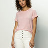 Person with curly hair in a "Perfect Tee Remix" by Sanctuary Clothing in earth pink, paired with white pants, standing against a plain background.