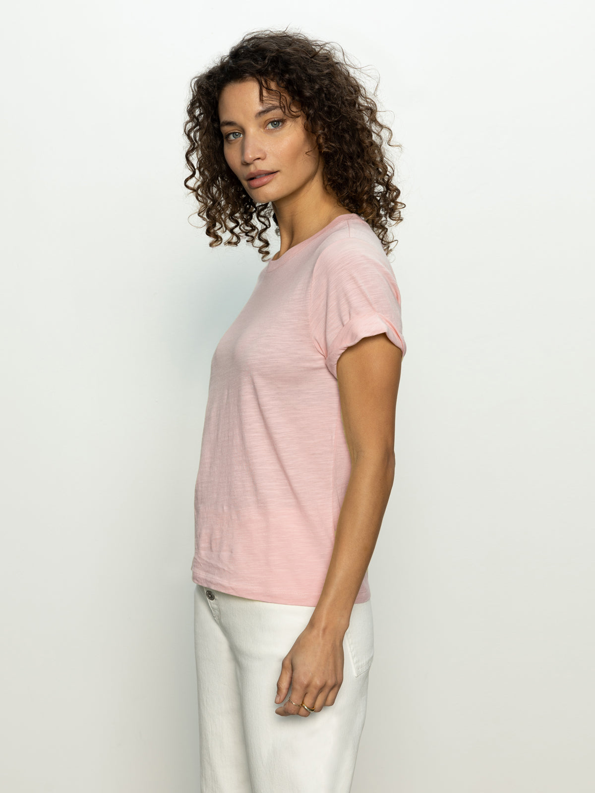 A person with curly hair stands sideways against a plain background, wearing Sanctuary Clothing's perfect tee remix in earth pink and white pants. They have a neutral expression with hands resting at their sides.