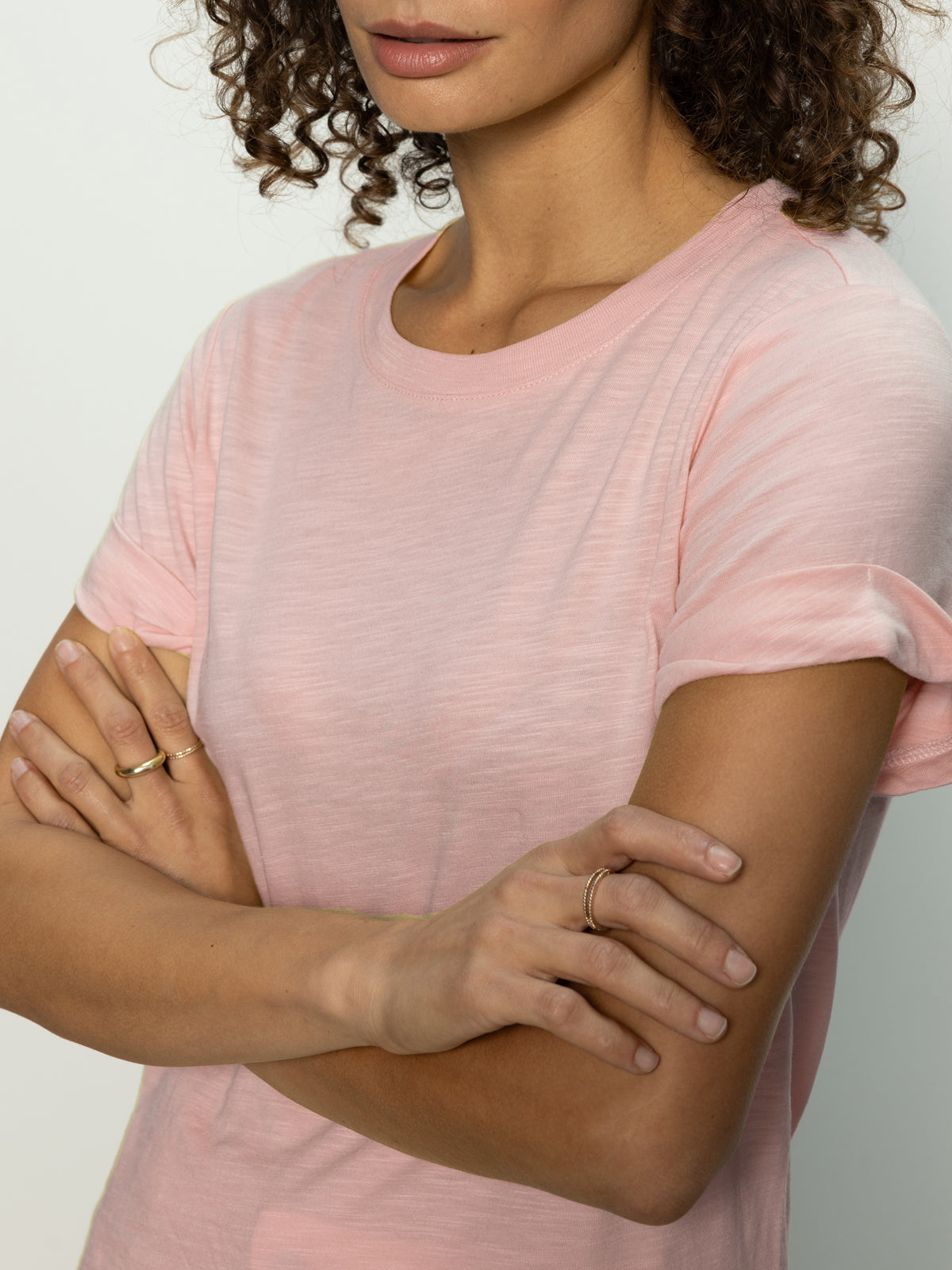 With a neutral expression, a person with curly hair stands with arms crossed, wearing Sanctuary Clothing's perfect tee remix in earth pink. Two rings can be seen on their fingers against the backdrop of a plain white wall.