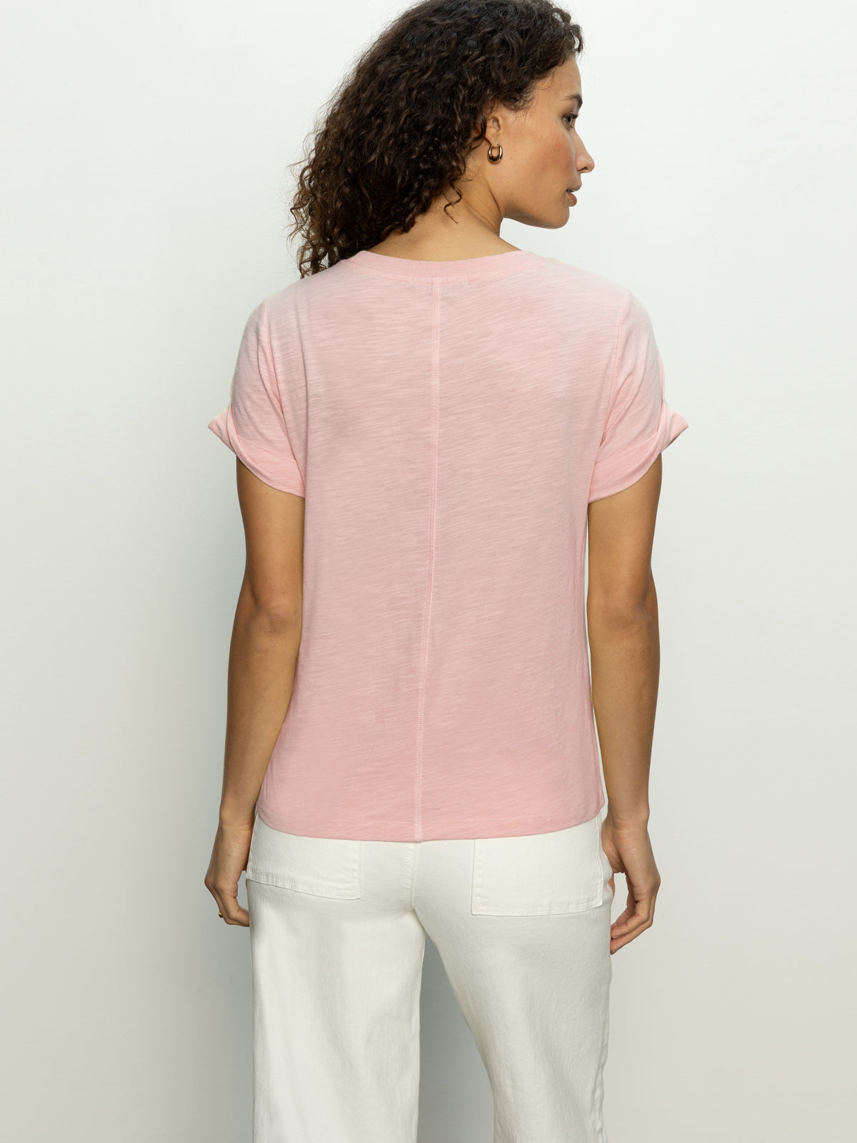 A person with curly hair, facing away against a plain background, is wearing a Sanctuary Clothing's perfect tee remix in earth pink paired with white pants.