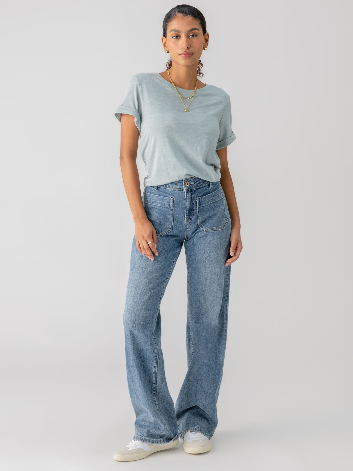 A woman stands confidently against a plain background, wearing the "Perfect Tee Remix Eucalyptus" by Sanctuary Clothing, paired with high-waisted blue jeans and white sneakers. Her curly hair is pulled back and she's accessorized with a gold necklace and earrings. Her left hand rests casually in her pocket.